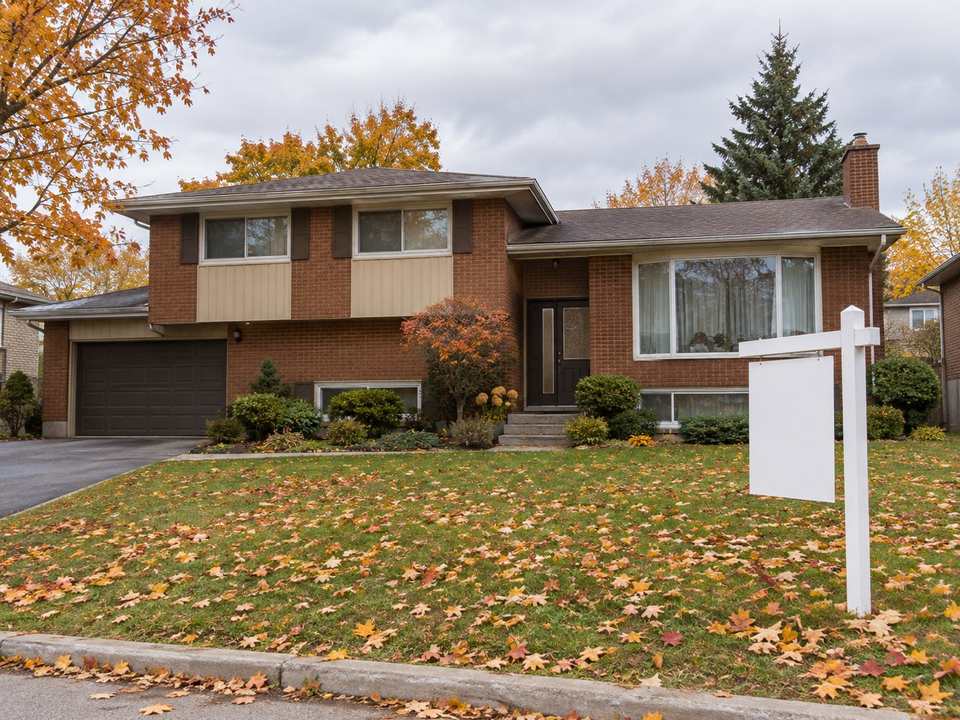 A 1970s brick backsplit house in autumn with a blank real estate sign on the front lawn